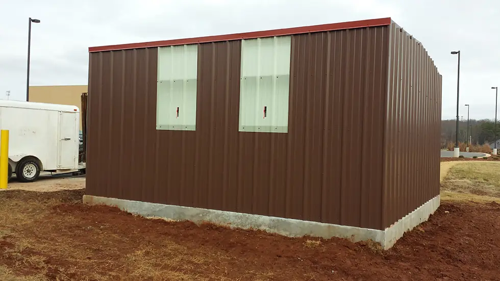 Brown-panel metal building with two windows and a white utility trailer parked in front under an overcast sky